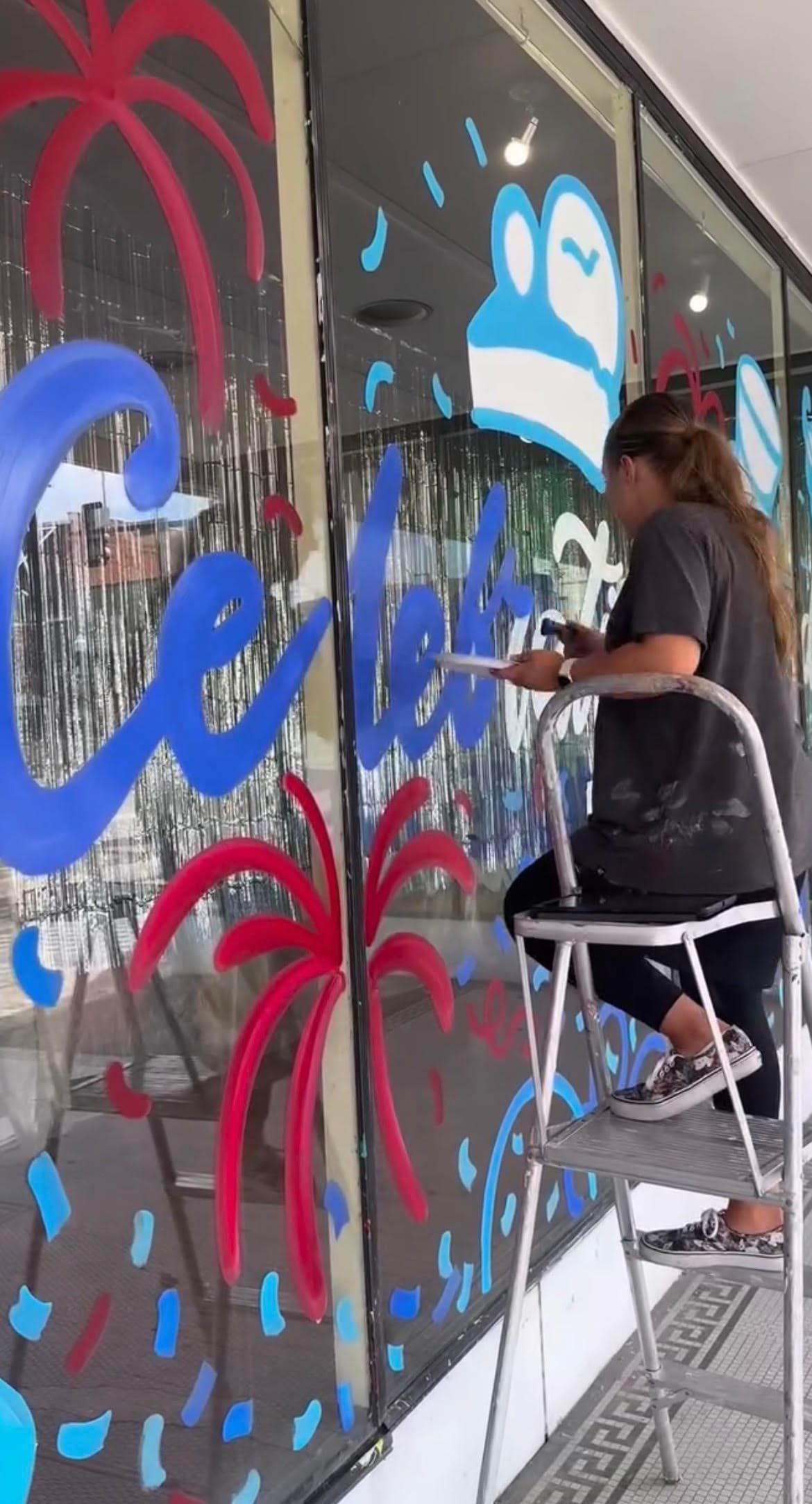 Woman on a step ladder paints festive red and blue designs on a glass storefront window.